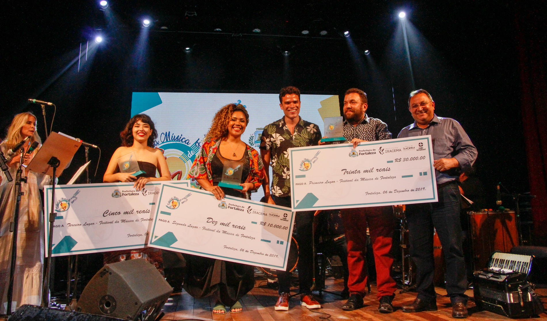 duas mulheres e dois homens em cima de palco segurando réplicas grandes de cheques e sorrindo